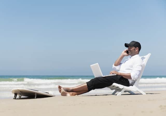 A Businessman Sits On A Beach Chair On The Beach Working On A Laptop Computer And Talking On The Phone With His Surfboard Sitting At His Feet; Tarifa Cadiz Andalusia Spain --- Image by © Ben Welsh/Design Pics/Corbis